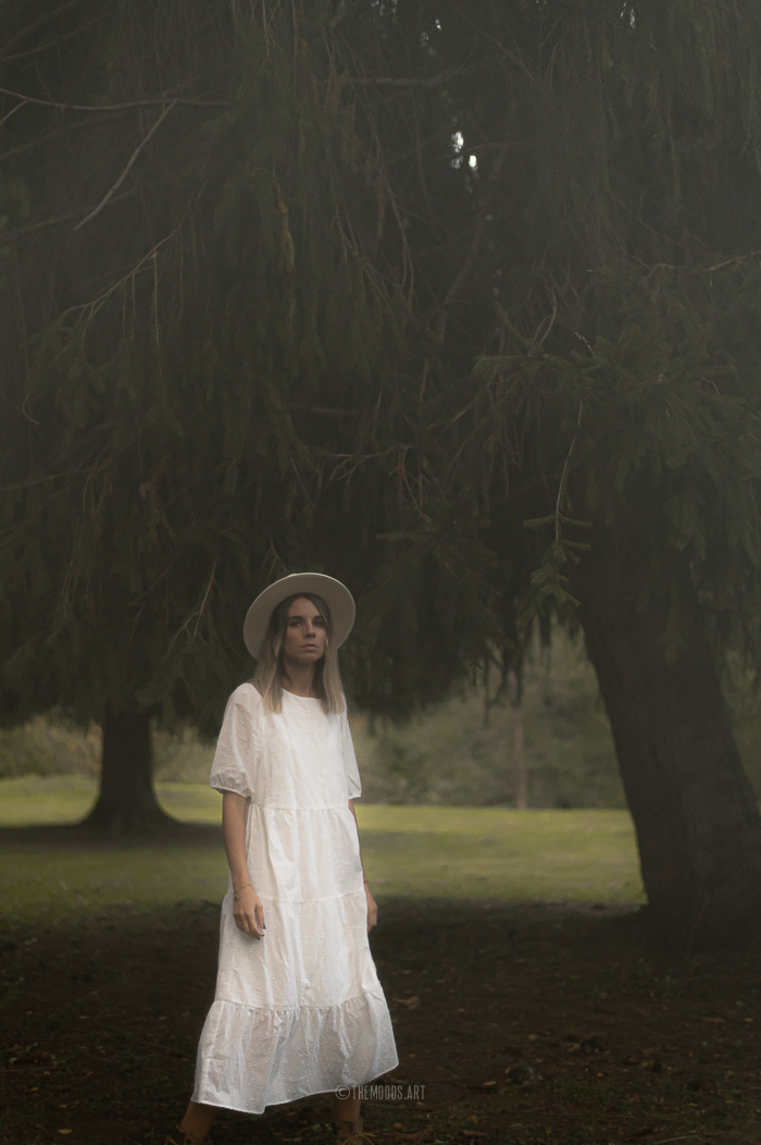 Photographie d'une femme avec une robe blanche au milieu des sapins le soir d'Halloween.